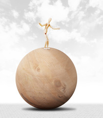 Man keeps balance standing on the top of a huge wooden ball. Abstract image with a wooden puppet