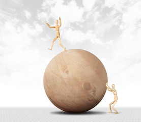Man pushes a huge wooden ball and another tries not to fall from it. Abstract image with a wooden puppet