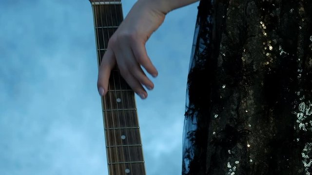 A girl in a concert dress approaches the guitar neck and gently passes her hand over the strings. Close-up of female fingers. Clubs of smoke in the background. Theme for the music video.