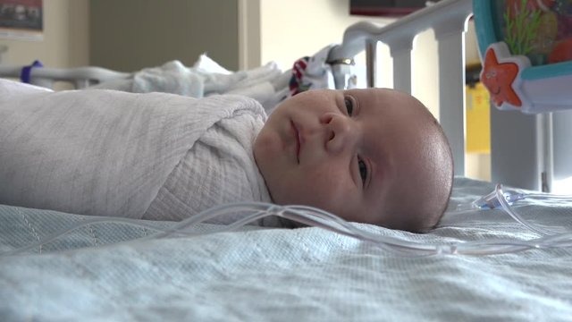 A baby is laying swaddled in a hospital crib attached to an IV line.  Shot in high speed photography.