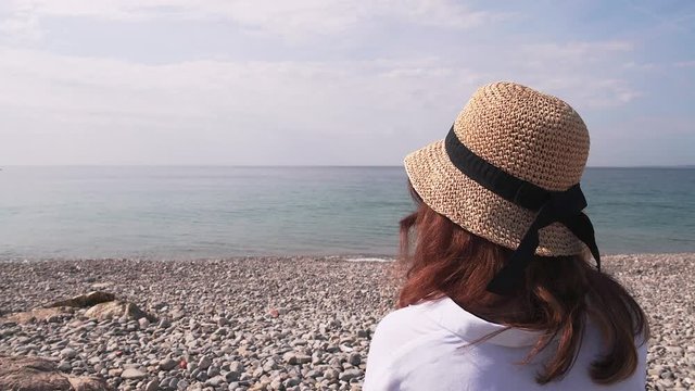 Unrecognizable young woman with long brown hair wearing a hat and a white shirt sitting on pebble beach and looking at sea. Locked down real time medium shot
