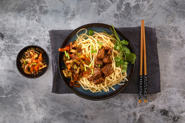 Chinese noodles with grilled meat, vegetables and greens on a gray table. Top view
