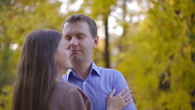 adult funny man is hugging his pretty wife in autumn park, snuggling to her face and bitting nose jokingly
