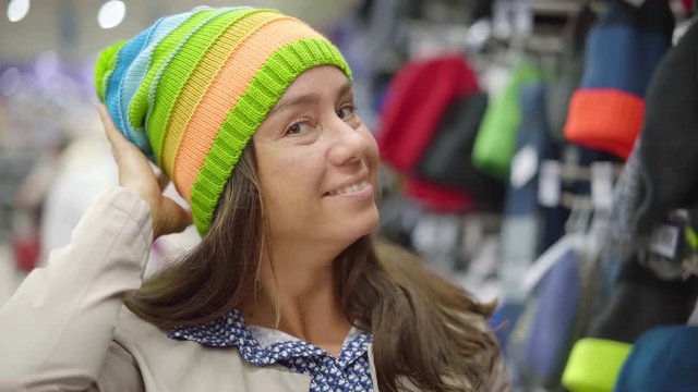 smiling female shopper is demonstrating bright knitted hat on her head in a hall of clothing store
