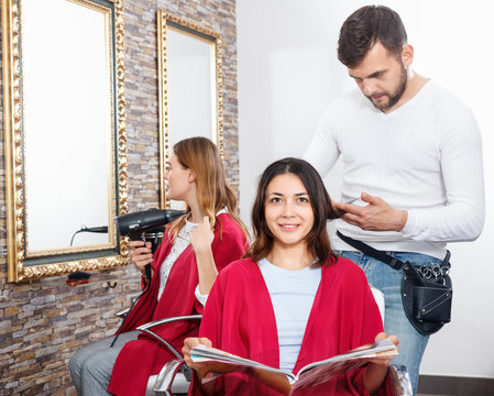 Positive young man hairdresser cuts hair of young woman with magazine at salon