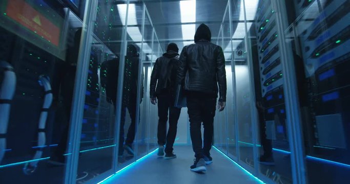 Wide shot from behind of a two hackers walking through corporate data center with rows of working rack servers