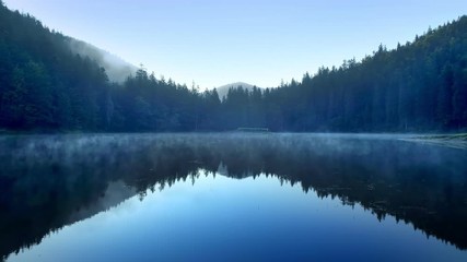 Clear blue mountain lake covered with low fog Aerial flight from water surface level Reflections of sky and mountains falling in water