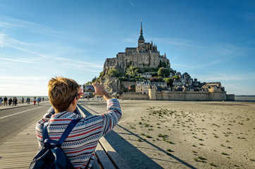 A tourist takes photos of Mont Saint Michel with her smartphone - obrazy, fototapety, plakaty