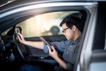 Young Asian businessman with glasses reading news on digital tablet while sitting on driver seat in his car. Business and technology concept