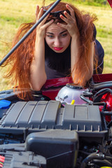 Fototapeta na wymiar A young woman stands by the car with a raised hood and puzzled, looking at the engine of the car. Car breakdown on the road