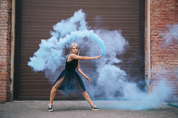 attractive girl looking at camera and dancing in blue smoke on street