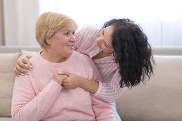 Portrait of old mother and mature daughter hugging at home. Happy senior mother and adult daughter embracing with love on sofa. Cheerful woman hugging from behind older mom and looking at each other.