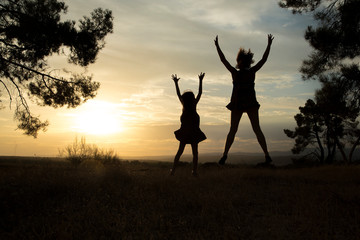 Backlight of a mother and her daughter jumping in a pine forest with yellow sky