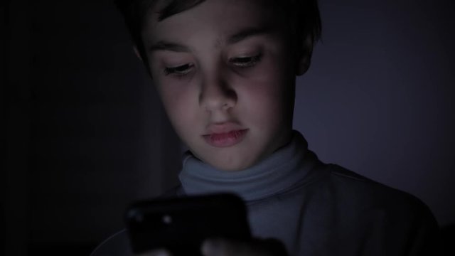 Happy Boy Enthusiastically Plays on a Smartphone in a Game in the Dark at Home. The face of the child is lit by a bright monitor