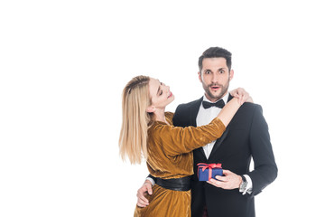 attractive woman hugging shocked boyfriend in suit with wrapped gift isolated on white