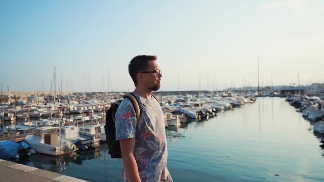 Cheerful bearded man is strolling near pearce with docked boats in sunny morning. Traveler is exploring landmarks and beautiful places of sea city