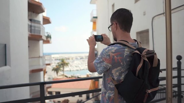 Young man is photographing and filming by smartphone a picturesque view. He is standing on open terrace in sunny summer day and looking on sea port
