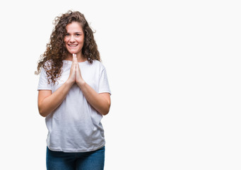 Beautiful brunette curly hair young girl wearing casual t-shirt over isolated background praying with hands together asking for forgiveness smiling confident.