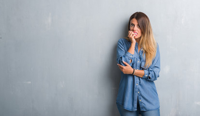 Young adult woman over grunge grey wall wearing denim outfit looking stressed and nervous with hands on mouth biting nails. Anxiety problem.