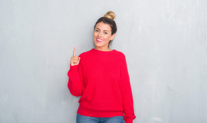 Obraz na płótnie Canvas Young adult woman over grey grunge wall wearing winter outfit showing and pointing up with finger number one while smiling confident and happy.
