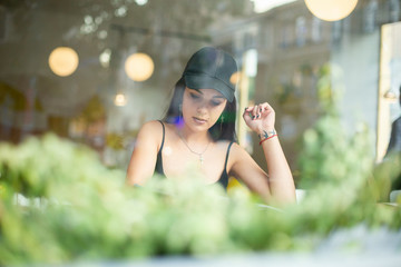 Portrait of a young beautiful girl sitting alone in a cafe and reading a fashion magazine. Shot through the window.