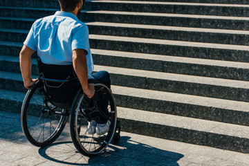 back view of disabled man using wheelchair on street and stopping near stairs without ramp