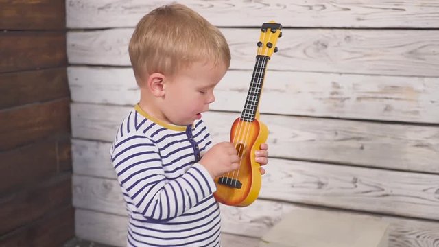 happy one year old boy plays his guitar or ukulele and sings songs, sitting in the wooden room.