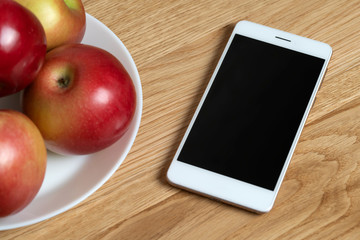White smartphone with a blank screen on a wooden table near a plate with red apples.