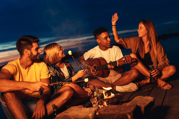 Group of young friends enjoying at the lake at night. They sitting around the fire singing and having fun at camping.