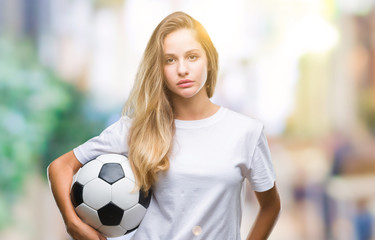 Young beautiful blonde woman holding soccer ball over isolated background with a confident expression on smart face thinking serious