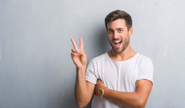 Handsome young man over grey grunge wall smiling with happy face winking at the camera doing victory sign. Number two.