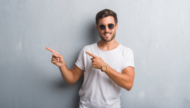 Handsome young man over grey grunge wall wearing sunglasses smiling and looking at the camera pointing with two hands and fingers to the side.