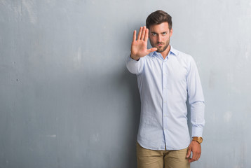 Handsome young business man over grey grunge wall wearing elegant shirt doing stop sing with palm of the hand. Warning expression with negative and serious gesture on the face.