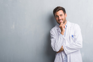 Handsome young professional man over grey grunge wall wearing white coat looking confident at the camera with smile with crossed arms and hand raised on chin. Thinking positive.