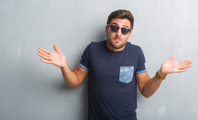 Fototapeta premium Handsome young man over grey grunge wall wearing sunglasses clueless and confused expression with arms and hands raised. Doubt concept.