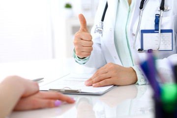 Doctor showing Ok sign with thumb up to patient while sitting at the desk in hospital office, close-up of human hands. Medicine and health care concept