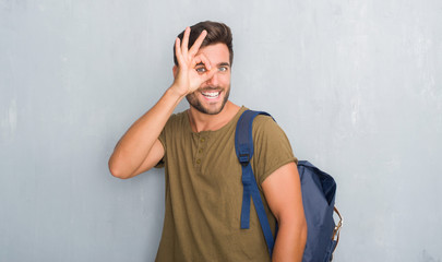 Handsome tourist young man over grey grunge wall wearing backpack doing ok gesture with hand smiling, eye looking through fingers with happy face.