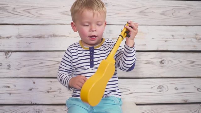 happy one year old boy plays his guitar or ukulele and sings songs, sitting in the wooden room.