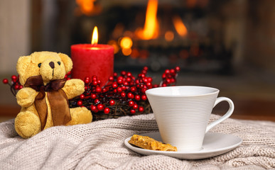 Cup of hot drink teddy bear candle in red Christmas decoration on cozy knitted plaid in front of warm fireplace. Cozy relaxed magical atmosphere in home interior. Holiday Christmas New Year concept.