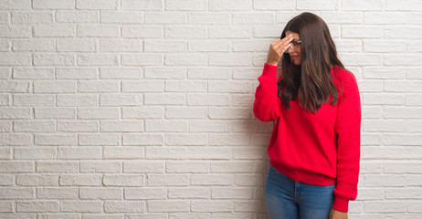 Young brunette woman standing over white brick wall tired rubbing nose and eyes feeling fatigue and headache. Stress and frustration concept.