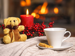 Cup of hot drink teddy bear candle in red Christmas decoration on cozy knitted plaid in front of warm fireplace. Cozy relaxed magical atmosphere in home interior. Holiday Christmas New Year concept.