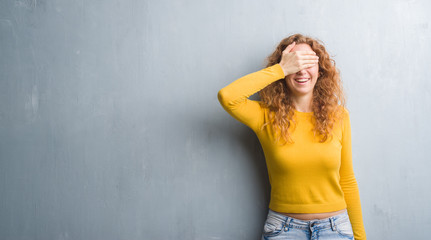 Young redhead woman over grey grunge wall smiling and laughing with hand on face covering eyes for surprise. Blind concept.