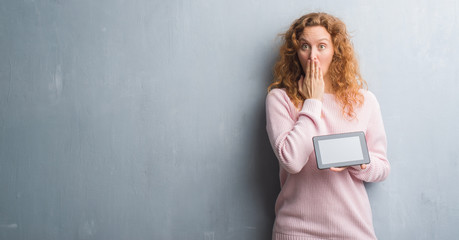 Young redhead woman over grey grunge wall using tablet cover mouth with hand shocked with shame for mistake, expression of fear, scared in silence, secret concept