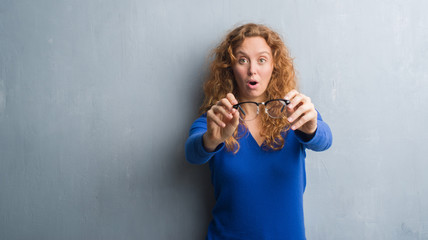 Young redhead woman over grey grunge wall holding optical glasses scared in shock with a surprise face, afraid and excited with fear expression