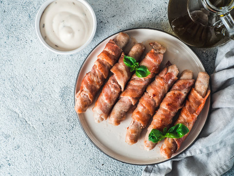 Ready-to-eat pigs sausages wrapped in bacon on plate. Fried savory sausages wrapped in bacon served fresh green basil leaves with sauce on background. Copy space for text. Top view or flat lay.