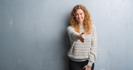 Young redhead woman over grey grunge wall smiling friendly offering handshake as greeting and welcoming. Successful business.