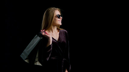 Woman in cool sunglasses and black dress, holding black shopping bag isolated on dark background in black friday holiday or christmas