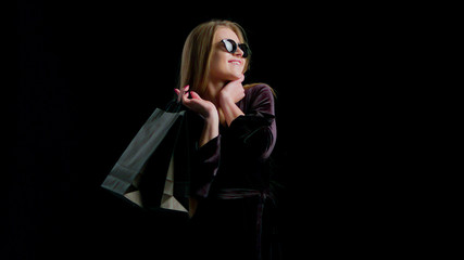 Woman in cool sunglasses and black dress, holding black shopping bag isolated on dark background in...
