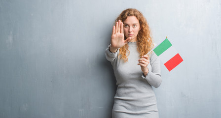 Obraz premium Young redhead woman over grey grunge wall holding flag of Italy with open hand doing stop sign with serious and confident expression, defense gesture