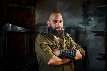 Brutal bearded blacksmith worker smokes after hard work. The man is tired, but smiling, his face is covered in mud and sweat